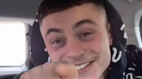 Family handout A young man is sat in a car pointing at the camera. It's a close-up of his head and he is smiling. He is wearing a black hoodie with white U-shaped squiggles on it.