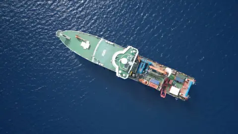 Kevin Church/BBC An aerial view of the RRS Discovery in the water of Santorini's caldera. At the front is the green deck and at the back are orange and blue shipping containers on deck.