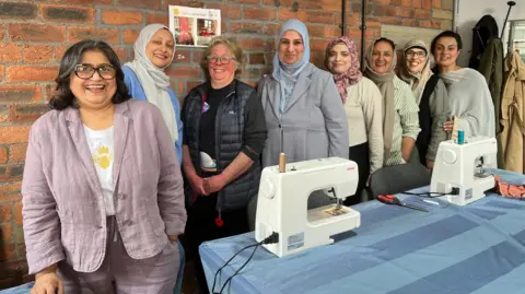 Aisha Iqbal/BBC A group of smiling women stand in front of a table with two s machines on it, as well as scissors and other tailoring equipment