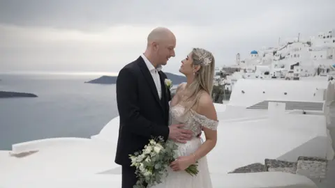 Kevin Church/BBC A photograph of Tom and Kristina wearing bridal outfits standing on the white rooftops of Santorini, facing each other in an embrace. Kristina is holding a bouquet of flowers.