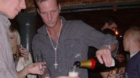 UGC Adam Minett stands in a nightclub in Liverpool, pouring champagne from a bottle in his left hand. He is surrounded by other people. 