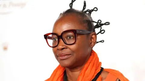PMC/Getty Images Koyo Kouoh wears thick rimmed glasses, an orange scarf and an orange top.
