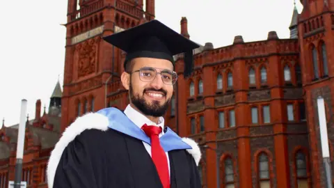 Hamza Al-Huseini Hamza Al-Huseini stands outside the University of Liverpool's Victoria Building in his graduation cap and gown. He wears glasses and has a short beard.