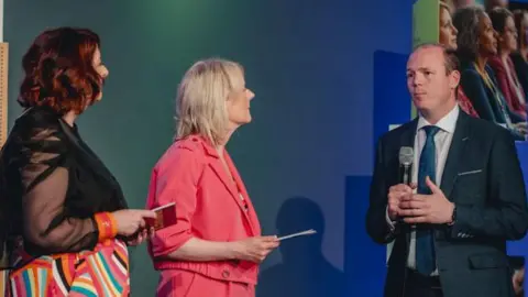 Rebootandreturn Three people on a stage. A woman on the left has shoulder length red hair. She is looking away from the camera. She is wearing a black mesh shirt and colourful stripy trousers. In the middle a woman with blonde hair is looking at the minister. She is wearing a pink matching suit. On the right, the minister is wearing a grey suit and a blue tie. He has short brown hair and is holding a microphone.