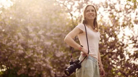 Bethany Clarke Bethany Clarke, a woman with long brown hair wearing a pink t shirt and trousers, holds her camera as she stands beneath a cherry blossom tree 