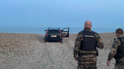 Marianne Baisnée/BBC Two police officers walk towards an abandoned black car sitting on a shingled beach, one door still open