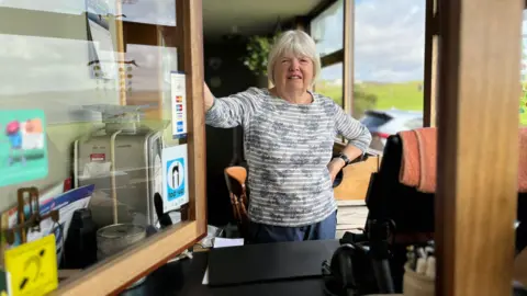 BBC Woman in a blue stripy top stands next to an open hatch/window. A set of scales is on one side and the green countryside can be seen through a window behind her. 