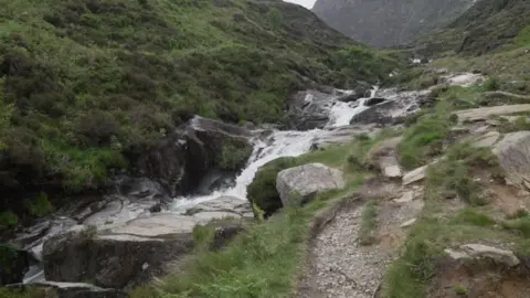 BBC A mountainside with greenery on either side and water running down the middle in a stream with lots of boulders and rocks