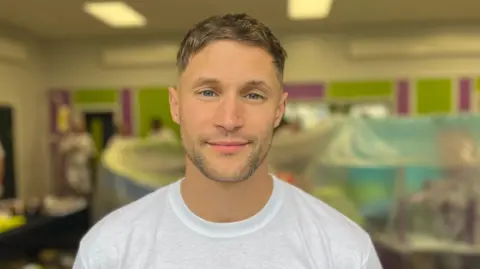 Marco is a man aged in his 30s. He is wearing a white t-shirt and has dark blonde hair and blue eyes. He is smiling with his lips pursed.