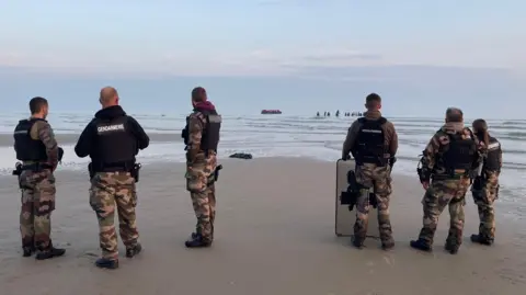 Marianne Baisnée/BBC Six French gendarmes stand facing the sea, their backs to the camera. In the distance you can see a large inflatable boat close to where dozens of people are gathered in waist-deep water