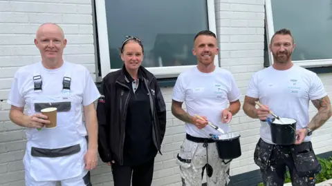 A woman with dark hair tied back, wearing a black Crown Paint jacket, and black trousers stands between three men wearing white t-shirts and paint-covered trousers, holding paint brushes.