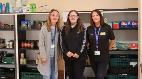 North Tyneside Council Rachel Fairless is standing in the middle of the pantry with two other women. Behind them are shelves filled with pantry food and tins.