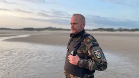 Marianne Baisnée/BBC A man in his fifties dressed in police uniform, with a bullet-proof vest, stands on the sand of a beach looking out to see