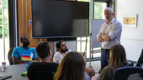 Caroline Sedilekova A man with white hair, wearing a white shirt, standing with his arms folded. He is addressing five men and women gathered around a table, three of whom have their backs to the camera but are looking towards him. They appear to be in a meeting room, with a central table, large-screen TV and whiteboard. Windows look out onto bushes.