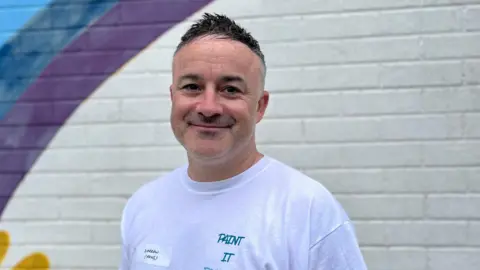 Darren has dark hair with grey at the sides. He is smiling at the camera with his mouth closed and wearing a white t-shirt with a name badge.