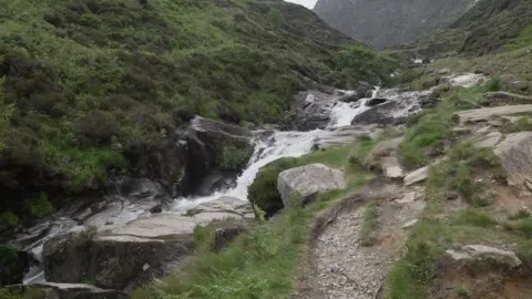 BBC A stream cascades down a series of rocks alongside a walking path found on the Watkin Path near the summit of Yr Wyddfa.. The bank slopes upwards on the left hand side.