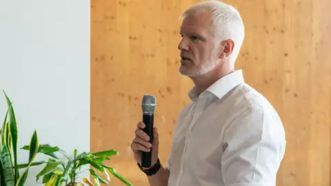 Caroline Sedilekova A side profile of a man with cropped white hair and close-cut beard addressing a room, holding a microphone. The man is wearing a white shirt and standing by a wooden panel background.