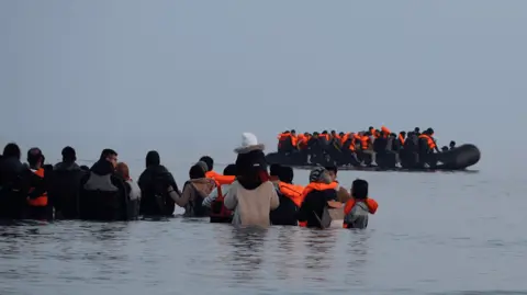 Lea Guedj/BBC A group of people stand in waist-deep water with an overloaded inflatable boat in front of them