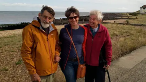 Christine Bell Colin, Christine and Beryl Bell are standing in front of a beach. Colin - an older, bearded man on the left of the three is smiling. He's wearing an orange raincoat. Christine, in the middle of the three, is wearing a blue coat and sunglasses. She has short brown hair. On the right, Beryl is an older woman in a red fleece coat. She is smiling at Christine and holding onto a walking stick.