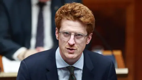 PA Media O'Toole is speaking in the assembly. He is wearing a navy suit and white shirt. He has ginger hair and wears round clear framed glasses.
