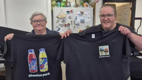 Two women hold up t-shirts they have printed. One has drinks bottles with rock guitar branding and the other shows figures in bucket hats and sunglasses. Both shirts feature Heaton Park branding.