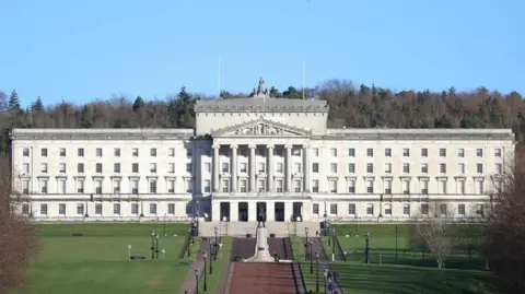 PA Media Stormont is a large stone building with stairs leading up to it. It is rectangular shape and has four rows of windows along the front, and a stone turrets at the front. On top of the building is a metal statue. Dotted around the grounds are black lampposts and benches for people to sit on. There is a statue feature in front of the steps to the front door.