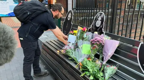 BBC A man with dark hair touches tributes on the bench.