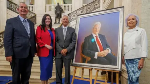PA Media A group picture of Poots, left, is smiling at the camera. He is wearing a dark grey suit and light blue shirt. He has combed back grey hair. Little-Pengelly is next in the line. She is wearing a bright red blazer and blue knee length dress. She is smiling at the camera and has long brown hair. Peter Robinson is standing next to the portrait which is in a silver frame. In the portrait he is wearing a dark coloured suit and red tie, he is sitting down and has white hair. Standing next to it he is wearing a grey suit and tie and has short white hair. On the right, Iris Robinson is smiling beside the portrait. She is wearing a white top which is asymmetrical, and a blue skirt. She has slightly purple short hair. 