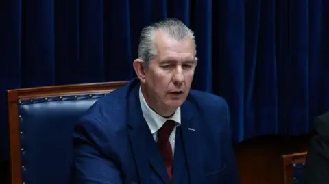 PA Media Edwin Poots is sitting in the Speakers chair. It is navy blue with a wooden frame. Poots is wearing a navy blue suit and red tie, with a white shirt. He has combed back grey hair.