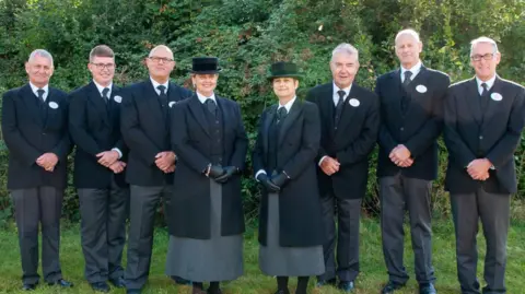 Susan Whymark The Susan Whymark Funeral Directors team stand in a line together in their uniform and smile at the camera. They are all wearing black suits and two women at the front where grey shirts with black hats.