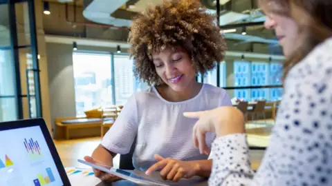 AFP via Getty Images Two women sit opposite each other looking at a laptop screen and an iPad device. They are in an open-plan office with windows behind them as well as chairs and a desk.