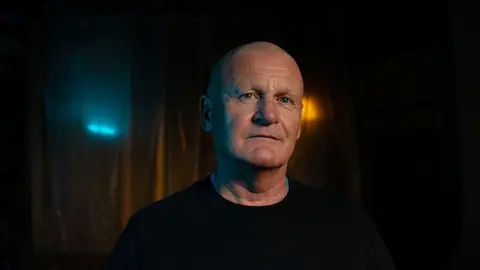 BBC/Double Act Productions Andrew Dodd has a bald head and blue eyes and is lightly tanned. He is approximately in his late 50s. He is looking at the camera. He wears a plain black jumper. The background is dimly lit with lights behind a translucent curtain.