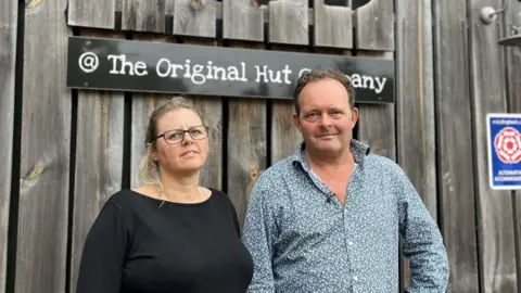 Chrissie Riedy A man and woman stand outside a cafe. She has blonde hair in a ponytail and is wearing a black top and black glasses. He has brown hair with a blue floral shirt on. There is fencing behind them, with a sign saying The Original Hut Company
