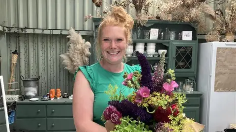 BBC/Jadzia Samuel A woman smiles widely holding a bouquet of flowers.