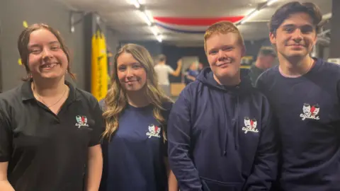 BBC Two teen girls and two boys wearing tops with a theatre group logo standing next to each other while a rehearsal continues behind them. The girl on the left has a blue t-shirt and is smiling, her brown hair is tied but she has two strands of hair down the left and right side of her face. The girls next to her has long blonde hair with brown streaks and has a blue t-shirt on. The boy to her right has ginger hair and a blue hoodie on, and the boy to his right has a blue t-shirt on and short dark brown hair. 