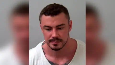 North Wales Police Terence Murrell, has cropped dark hair a light beard around the edge of his face and moustache. His skin is quite red or tanned, and he wears a white Tshirt. The edge of an extensive tattoo can be seen under his shirt. He is looking down. The picture is a police mugshot from his arrest. The background is grey.