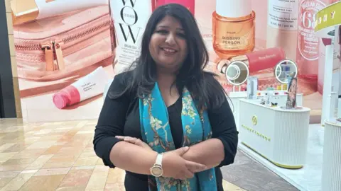 A woman with long black hair smiling at the camera, stood in front of a beauty store with hoarding. The hoarding has large makeup icons on it. 