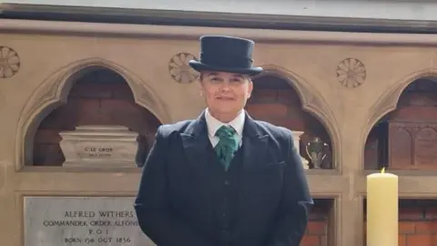 Susan Whymark Ms Whymark smiles at the camera while she stands inside a church. She wears a smart black coat, waistcoat, white shirt, green tie and black hat.