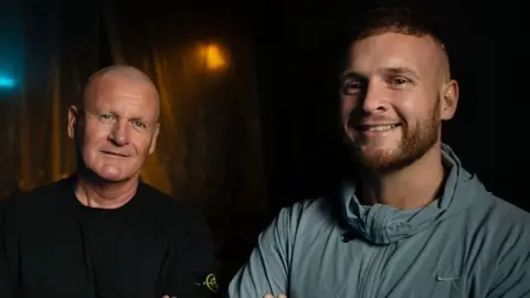BBC/Double Act Productions Andrew and Macaulay Dodd pictured standing next to each other. Both looks at the camera and smiles. Andrew wears a black top and Macaulay a light blue sports zip-up top.