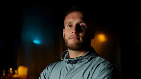 BBC/Double Act Productions Macaulay Dodd has short brown hair and a beard and is lightly tanned. He is late 20s/early 30s. He is wearing a Nike branded zip up blue-grey sports top. He is looking at the camera but not smiling. The background is dark with a few dim downlights.