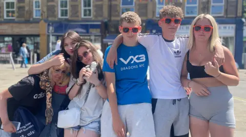 Six young people huddled together, divided into a trio of girls and two boys and a girl who are wearing orange-rimmed sunglasses. The girl on the right has blonde hair, grey shorts and a back crop top. The boy next to her has his arm around her, and has short curly light-brown hair and a white Adidas t-shirt on and grey shorts. His other arm is around a boy with short light-brown hair and a blue t-shirt which has VX3 Dragons RTC in white writing and grey shorts. Next to him are three girls huddled together. The one closest to him has a white t-shirt and denim shorts with a white handbag and is holding a cuddly toy, she has shades on and shoulder-length brown hair. Next to her is a woman with dark brown hair, she has a t-shirt with Cartman from Southpark on her top, her arm is on the head of the girl to the left of her. She has blonde hair and black Adidas top with a blue bag. 