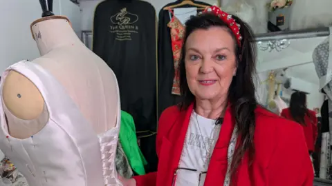 In this picture Angie is smiling at the camera as she stands next to a mannequin. Angie wears a red blazer and a white t-shirt. She's also wearing a sparkly red headband and has long black hair. 