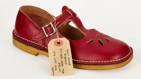 Shoemakers Museum Clarks leather children's shoe. The shoe is red with a small piece of cardboard and string attached.