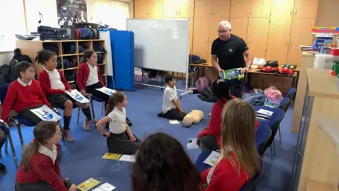 CHRIS MCHUGH/BBC Gary stands at one end of a classroom. Children are in front of him, some on chairs and some sat on the floor. He is holding a defibrillator and explaining how to use it.