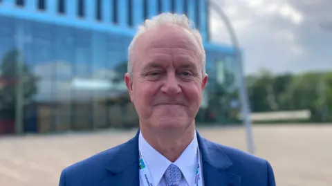Laura Coffey/BBC A man in a blue suit with a North Northamptonshire Council lanyard stands in front of the Corby Cube.