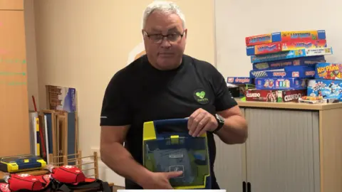 BBC Gary Elkins stands in a classroom holding up a mobile defibrillator. He has a black t-shirt and has short white hair with glasses. A stack of board games can be seen behind him and a table has further items used for his first aid course.