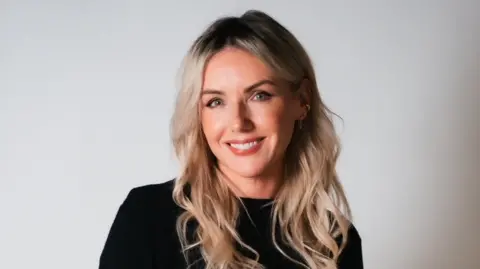 Dora Paphides Sali wears a black top and is smiling at the camera. It's a professional headshot. She has wavy blonde hair.
