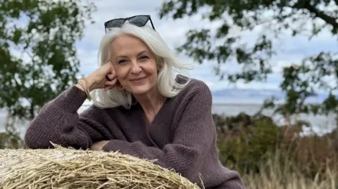 Valerie Mackay Valerie is outside, in a field, leaning on a bale of hay. She's wearing a purple/brown knit jumper and has black sunglasses perched on top of her head. She's smiling.
