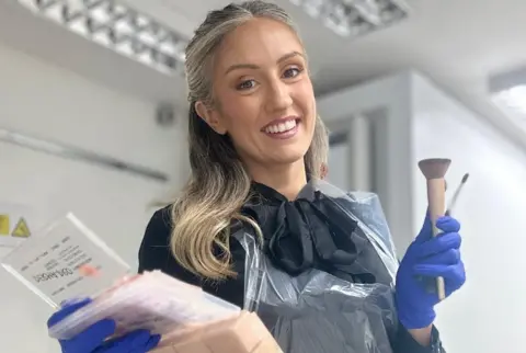 Collect/PA Real Life A woman with blonde hair is smiling at the camera. She is wearing a black top and blue gloves and is standing in front of a light-coloured wall.
