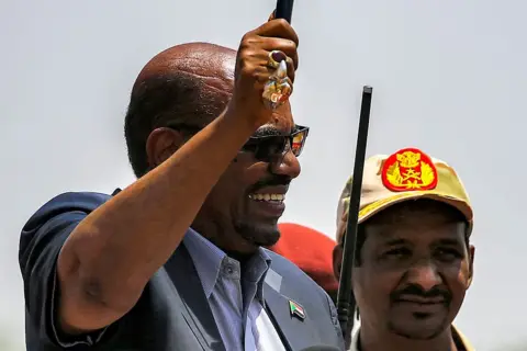 AFP via Getty Images Omar al-Bashir (L) waves a walking stick as he gives a speech at the headquarters of the Rapid Support Forces (RSF) paramilitaries in Umm al-Qura in South Darfur State, while RSF commander Mohamed Hamdan Daglo, wearing a cap, looks on